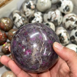 以下が含まれることがあります： 暗いグレーと紫の色合いが混ざり、鮮やかなピンク色の斑点がある、研磨された球形の宝石。 手に持たれており、背景には他の同様の石があります。