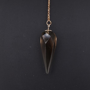 Może przedstawiać: Fasetowany wahadło w kształcie łzy, wykonane z ciemnego, półprzezroczystego materiału, zawieszone na złotym łańcuszku. Wahadło wisi na ciemnym tle, podkreślając jego geometryczny kształt i kolor.