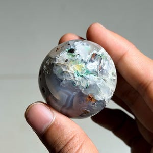 Könnte beinhalten: Ein polierter, kugelförmiger Edelstein mit einer Mischung aus grauen, weißen und blauen Bändern. Die Oberfläche weist grüne, braune und orangefarbene Flecken auf. Der Stein wird zwischen zwei Fingern gehalten, was seine natürlichen Farben und Texturen zeigt. Ein dekorativer Artikel.