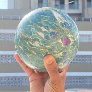 May include: A polished spherical stone featuring a marbled green base with prominent inclusions of deep red and dark blue mineral deposits throughout the surface, showcasing a smooth, glossy texture and a rounded, orb-like geometric shape.