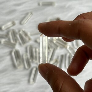 May include: A clear quartz crystal point, also known as a wand, is held between two fingers. The crystal is long and thin, with a pointed tip. The crystal is surrounded by other smaller quartz crystals.