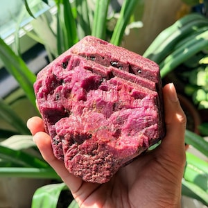 Könnte beinhalten: Ein großes, rohes Rubinkristall-Exemplar in tiefem Magenta und Karmesinrot. Der Stein weist eine hexagonale, geschichtete Struktur mit einer rauen, texturierten Oberfläche und kleinen, dunklen Einschlüssen in der Kristallformation auf.