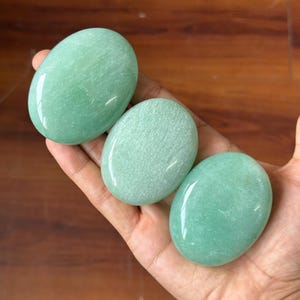 Può includere: Tre pietre verdi lisce e ovali sono tenute in una mano. Le pietre hanno un aspetto lucido e brillante e variano leggermente nella tonalità, da un verde più chiaro a uno più scuro. Lo sfondo è una superficie di legno.