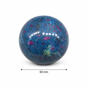 Puede incluir: Una piedra esférica pulida con un color base azul profundo, que presenta inclusiones dispersas de magenta vibrante y pequeñas manchas de depósitos minerales verdes. Un gráfico de flecha negra debajo de la esfera indica un diámetro de 60 mm.