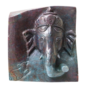 May include: A bronze-colored relief sculpture of the Hindu deity Ganesha, featuring an elephant head with detailed ornamentation. A small rat is depicted at the bottom left. The sculpture has a textured surface with hints of blue and green, set against a dark background.