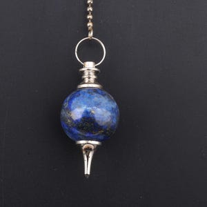 May include: A blue lapis lazuli pendulum with silver-colored metal accents. The pendulum has a spherical stone and a pointed tip, suspended by a chain. The background is a solid black.