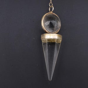 May include: A clear quartz crystal pendulum with a silver-coloured metal setting and chain. The pendulum has a rounded top and a pointed, cone-shaped bottom. The background is a solid black.