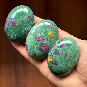 May include: Three oval-shaped green stones with red inclusions. The stones are smooth and polished, with a glossy finish. Each stone has a unique pattern of red spots and streaks. Two stones have small yellow stickers with numbers.