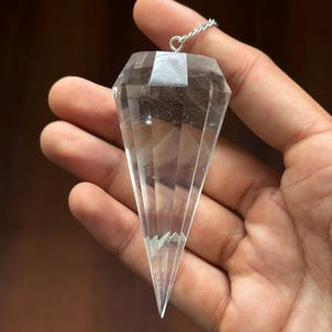 Könnte beinhalten: Ein klarer Kristallpendel mit facettierter, rautenförmiger Form. Der Kristall ist an einer kleinen Silberkette aufgehängt. Der Pendel wird in einer Hand vor einem verschwommenen braunen Hintergrund gehalten.