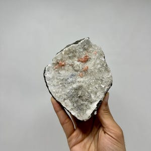 Puede incluir: Un espécimen mineral natural con un exterior oscuro y rugoso que encierra un interior cristalino. El interior es principalmente blanco con pequeños grupos dispersos de cristales de color rosa-naranja. El espécimen se sostiene en una mano sobre un fondo liso.