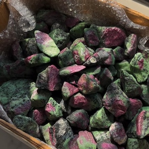 May include: A wooden box filled with raw ruby in zoisite stones. The stones are a mix of green, pink, and black, with a rough, natural texture. The stones are of various sizes and shapes.