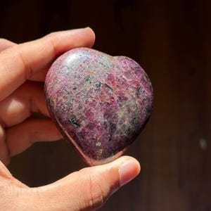 Könnte beinhalten: Ein herzförmiger Stein in tiefem Magenta, gesprenkelt mit dunkleren Flecken. Die polierte Oberfläche reflektiert das Licht und hebt die natürlichen Texturen und Variationen des Steins hervor. Ein einzigartiger Dekorationsartikel.
