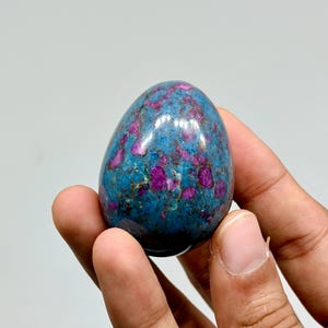 Op de afbeelding: Een eivormige gepolijste steen met een levendige blauwe basiskleur en ingebedde magenta en donkerroze minerale insluitingen over het gehele oppervlak. Het gladde, afgeronde object heeft een glanzende afwerking en een gevlekt uiterlijk.