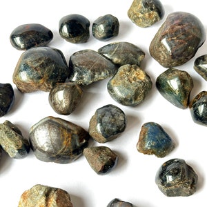 Puede incluir: Una colección de piedras de zafiro estrella azul y negro, algunas con un brillo metálico, sobre un fondo blanco.