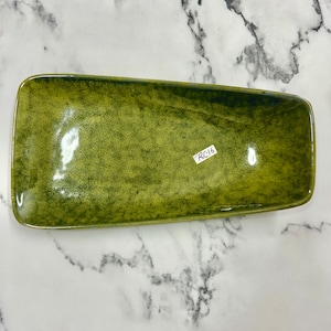 Peut inclure: Un plat de service rectangulaire en céramique vert olive aux coins arrondis. Le plat a une finition brillante et une texture mouchetée. Le texte "RC16" est visible. Idéal pour servir des amuse-gueules. Mesure environ 25 cm de long.