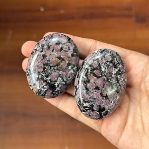 Puede incluir: Dos piedras ovaladas con un patrón moteado de color rosa intenso y negro. Las piedras son lisas y pulidas, con un acabado brillante. Las piedras se sostienen en una mano.