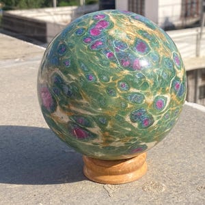 May include: A polished sphere made of green fuchsite stone with embedded magenta ruby crystals and golden mica inclusions. The sphere rests on a small, circular, light brown wooden stand, showcasing a smooth, rounded surface with natural mineral patterns.