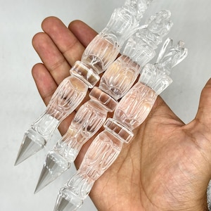 May include: Four clear, decorative crystal wands with pointed tips. The wands have a detailed, carved design and are held in a hand. The wands are made of a transparent material, reflecting light.