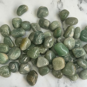 Puede incluir: Una colección de piedras lisas de aventurina verde. Las piedras son de varios tamaños y formas, con un color verde claro y un brillo sutil.