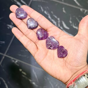 Op de afbeelding: Vijf kleine, gepolijste hartvormige stenen in verschillende tinten diep paars en magenta. Elke steen heeft een glad, reflecterend oppervlak met zichtbare interne insluitingen en natuurlijke aders, gerangschikt in een diagonale lijn.