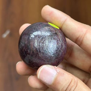May include: A polished, spherical gemstone with a deep burgundy color and swirling patterns. The orb is held in a hand, showcasing its smooth, reflective surface. A small yellow sticker is visible on the surface of the sphere.