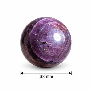 Peut inclure: Une pierre précieuse sphérique polie aux teintes magenta et violet profond, présentant des fractures internes naturelles et des textures cristallines. La sphère a un diamètre de 33 mm, comme indiqué par le graphique de mesure situé sous l'objet.