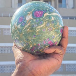 May include: A polished spherical stone featuring a mottled green base with embedded vibrant magenta and deep pink crystalline inclusions. The surface displays a smooth, glossy texture with swirling patterns of white and gold mineral veins throughout.