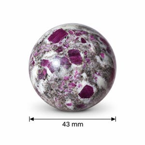Puede incluir: Una piedra esférica pulida con un patrón moteado de inclusiones minerales en color magenta intenso, blanco y gris oscuro. Una línea de medición negra con flechas indica un diámetro de 43 mm en el centro de la esfera.