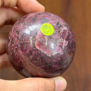 May include: A polished, spherical gemstone in shades of deep red and purple, held in a hand. A small, round sticker with the numbers "387" and the letters "GM" is affixed to the surface of the stone. The stone's surface has a textured appearance.