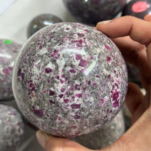 Könnte beinhalten: Eine polierte Kugel aus Rubin-Granit, die ein gesprenkeltes Muster aus tiefem Rosa und Grau aufweist. Die Oberfläche des Steins ist glatt, mit sichtbaren Rubin-Einschlüssen. Weitere ähnliche Kugeln sind im Hintergrund zu sehen.