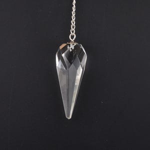 May include: A clear, faceted crystal pendulum suspended from a silver-colored chain. The crystal is elongated with a pointed tip and a geometric design. The background is a solid black.