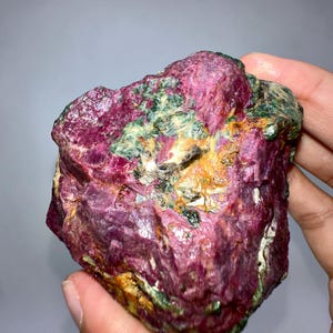 Puede incluir: Una gema de rubí en bruto, sin cortar, de color rojo intenso, acentuada por parches verdes y amarillos. La piedra natural tiene una superficie texturizada y una forma irregular, mostrando su belleza en bruto. Ideal para coleccionistas o para hacer joyas.