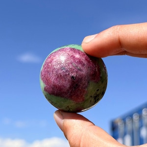 May include: A polished spherical gemstone featuring a vibrant magenta-red center surrounded by a mottled olive-green and black mineral matrix. The smooth, rounded surface displays natural color variations and crystalline textures throughout the stone.