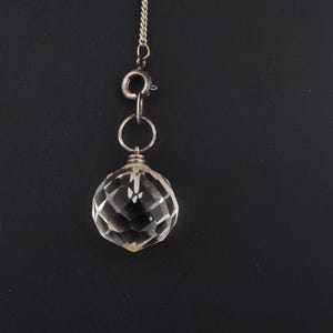 May include: A clear, faceted crystal pendant suspended from a delicate silver chain. The crystal is spherical with multiple facets, reflecting light. The chain has a small clasp. The background is a solid black.