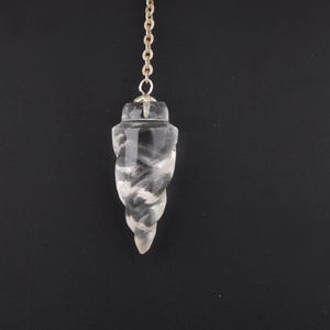 May include: A clear quartz crystal pendulum with a silver-colored chain. The pendulum is carved into a spiral shape. The background is a solid black.