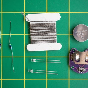 May include: A green cutting mat with various electronics components, including two small blue and black resistors, two clear LED lights, a spool of brown thread, a small purple circuit board with a battery holder, and a silver lithium cell battery labeled "Lithium Cell CR2032 3V SC".