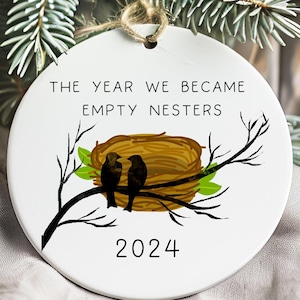 May include: White ceramic ornament with a brown nest and two black birds on a branch. The text "The year we became empty nesters 2024" is printed on the ornament.