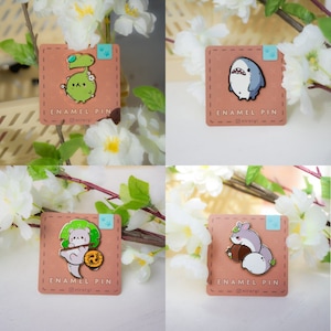 Könnte beinhalten: Vier Emaille-Pins mit niedlichen Comic-Tieren. Die Pins befinden sich auf einem braunen Hintergrund mit dem Text "ENAMEL PIN" und dem Social-Media-Handle "@miraryi". Die Pins sind von weißen Blüten umgeben.