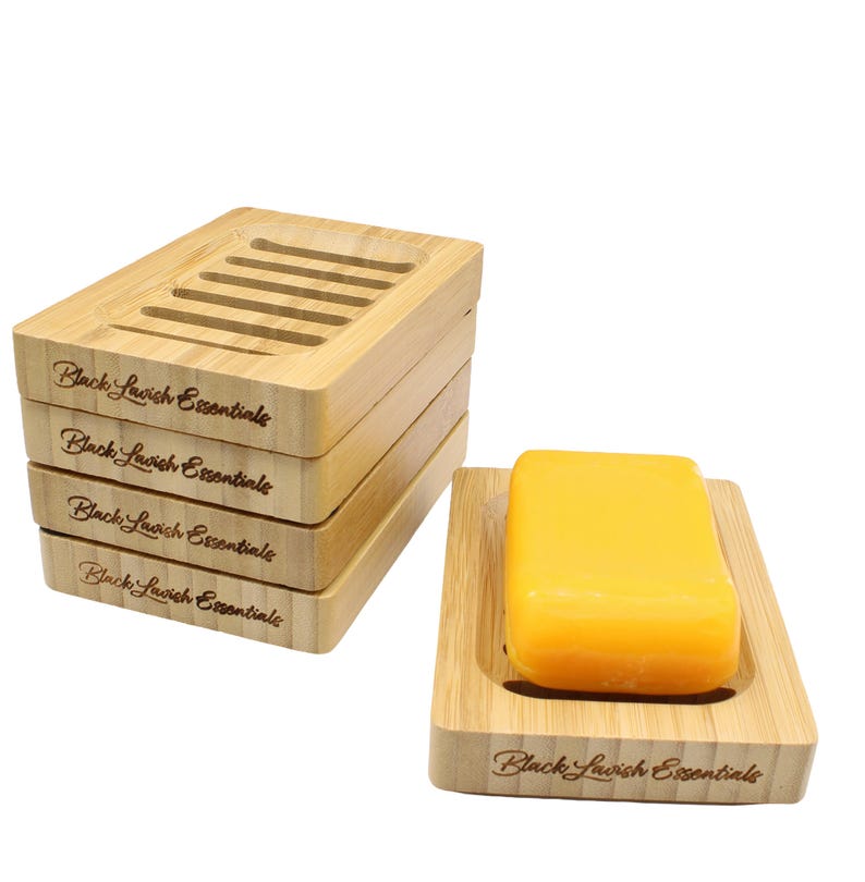 K&ouml;nnte beinhalten: Ein Set aus Bambus-Seifenschalen, darunter ein Stapel von vier und eine mit einem gelben Seifenst&uuml;ck. Jede Schale ist rechteckig und tr&auml;gt den Text "Black Lavish Essentials".