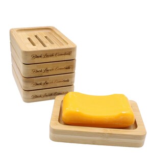 K&ouml;nnte beinhalten: Ein Set aus Bambus-Seifenschalen mit der Gravur "Black Lavish Essentials". Eine Schale enth&auml;lt ein gelbes Seifenst&uuml;ck. Die obere Schale hat drei Abfluss&ouml;ffnungen.