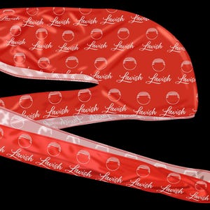 May include: A red satin durag with a white interior and trim. The durag is patterned with the word "Lavish" in a cursive font and a circular design. The fabric is folded and draped against a black background.