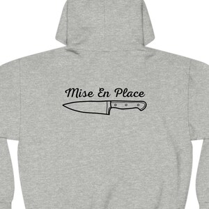 Puede incluir: Sudadera gris con capucha con el texto "Mise En Place" y un dibujo de un cuchillo de chef en la espalda.