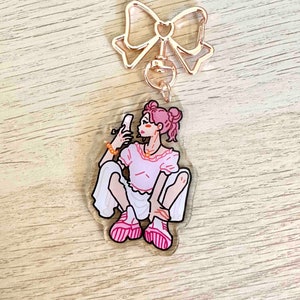 May include: A clear acrylic keyring with a cartoon illustration of a person with pink hair and a pink outfit taking a selfie. The keyring has a rose gold bow-shaped top and a lobster clasp.