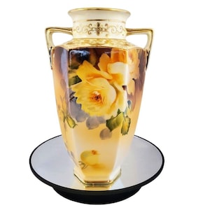 Puede incluir: Un jarrón vintage pintado a mano con un diseño floral. El jarrón presenta un borde y asas de color crema con detalles dorados. El cuerpo del jarrón está decorado con rosas amarillas y hojas verdes. El jarrón está sobre una superficie espejada.