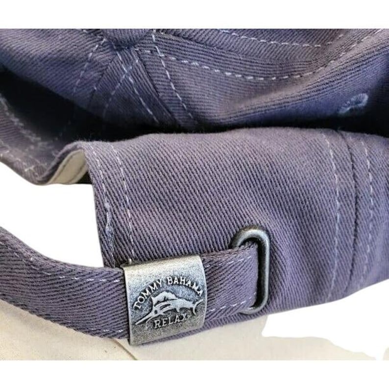 May include: Close-up of a dusty blue baseball cap. The cap features white stitching and a metal clasp with the Tommy Bahama Relax logo. The cap's brim and adjustable strap are visible.