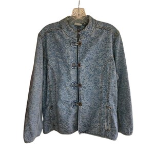 Puede incluir: Chaqueta vaquera azul claro con estampado floral. La chaqueta tiene cuello mandarín, mangas largas y tres cierres de palanca decorativos en la parte delantera. Se ven dos bolsillos. La chaqueta está colgada de una percha de plástico transparente.