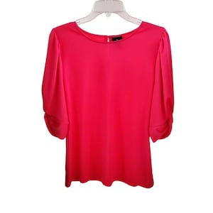 Könnte beinhalten: Eine leuchtend pinkfarbene Bluse mit kurzen Ärmeln, einem runden Ausschnitt und einer leicht gerafften Detail am Schulterbereich.