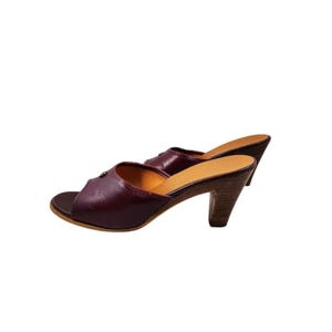 May include: A pair of burgundy leather slip-on mules with a peep-toe design. The shoes have a wooden heel, approximately 3 inches (7.6 cm) high. The shoes are set against a white background.