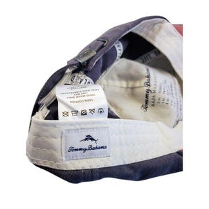 May include: A navy blue baseball cap with a white interior and a white label with the text "Tommy Bahama". The cap is made of 100% cotton and has care instructions printed on a tag.