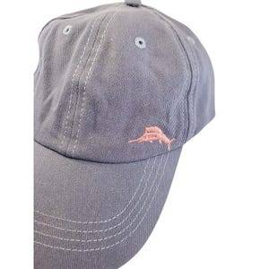 May include: A gray baseball cap with a curved brim and a small, pink embroidered fish logo on the front. The cap has a six-panel design with white stitching and a small button on top. The hat is made of a soft, durable fabric.